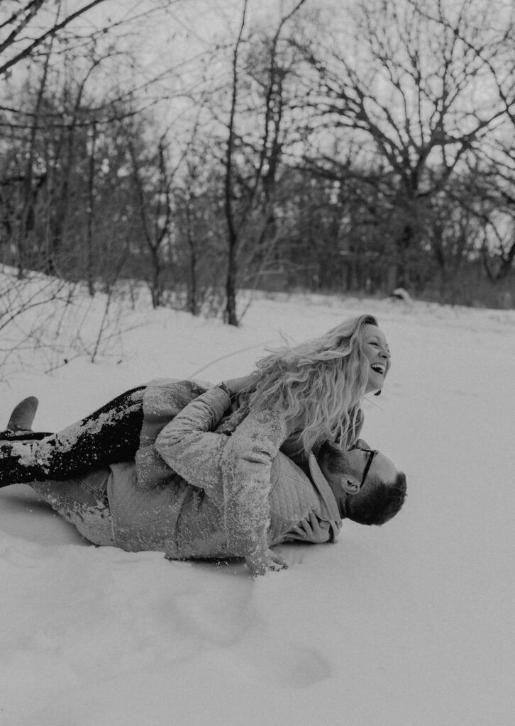 a black and white photo of a couple rolling around in the snow with the woman on top of the man laughing showing real emotion