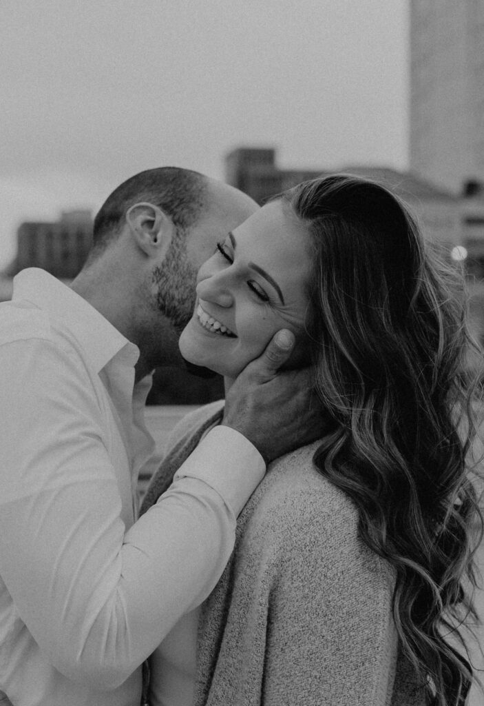 a couple laughs together on a rooftop while the man kisses the woman's cheek